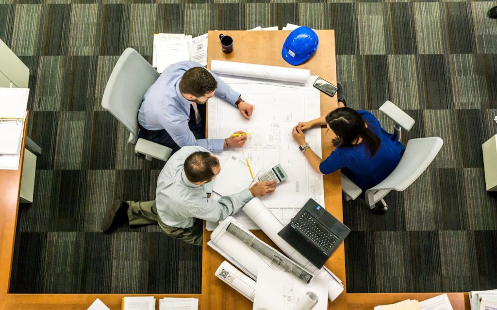 three-people-sitting-beside-table-416405 Top view of a team working on construction plans in an office setting.